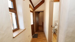 Ein schmaler Korridor mit Holzdielenboden, einem Fenster links und einer Tür, die in einen Raum mit einem Bett führt.