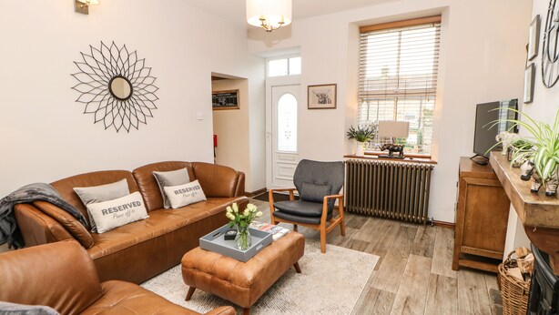 A living room with a brown leather sofa, a grey armchair, and a coffee table with a vase of flowers. There is a radiator, a television, and a window with blinds. - Magnolia Cottage (Richmond)