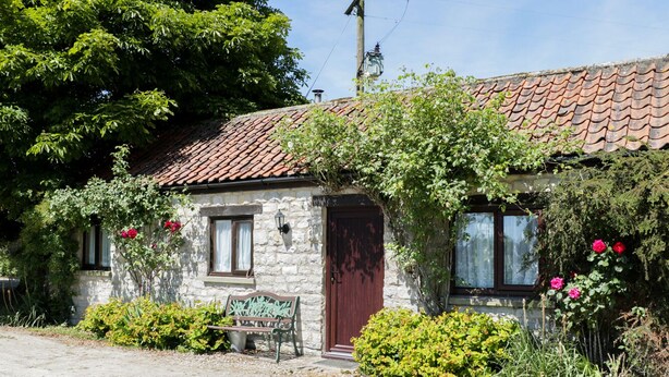 A stone cottage with a tiled roof, a wooden door, and a bench outside. - Rose Cottage (York)