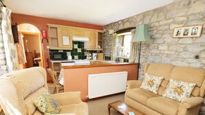 A cozy living room with a sofa, armchair, and coffee table. A kitchen with a stone wall and wooden cabinets is visible in the background. - Rose Cottage (York)