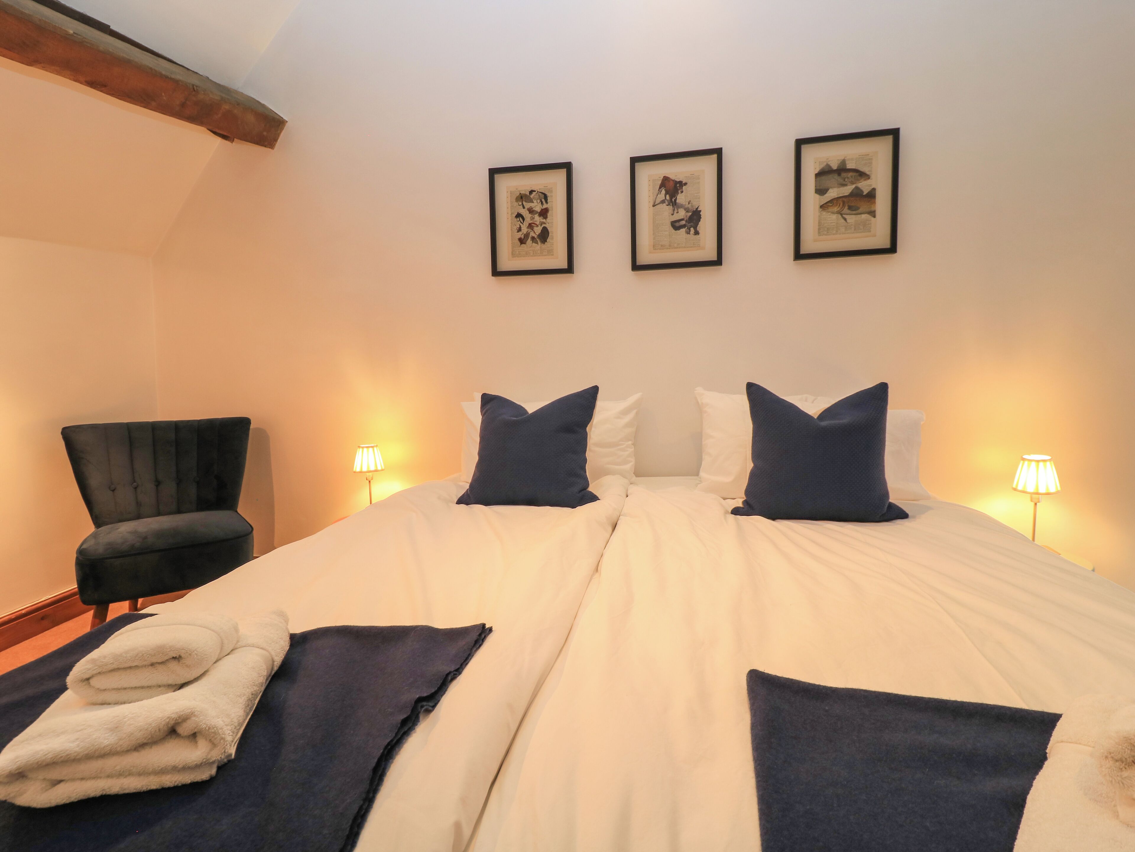 A bedroom with a large bed, two bedside lamps, a chair, and three framed pictures on the wall.