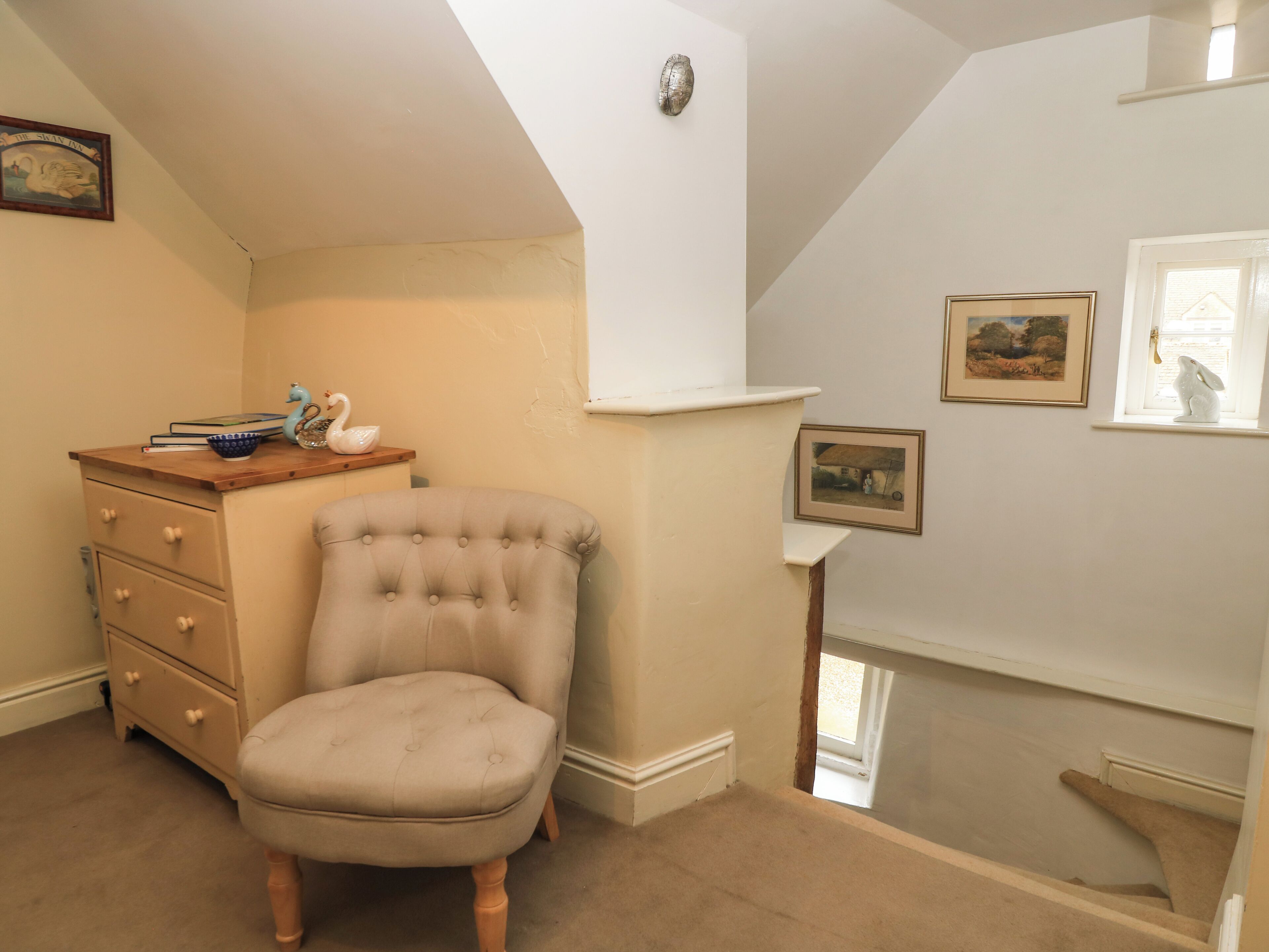 Une pièce comprenant un fauteuil à capitons, une commode en bois et une petite table agrémentée d’objets décoratifs. Des tableaux encadrés ornent le mur, et une fenêtre offre une vue agréable.