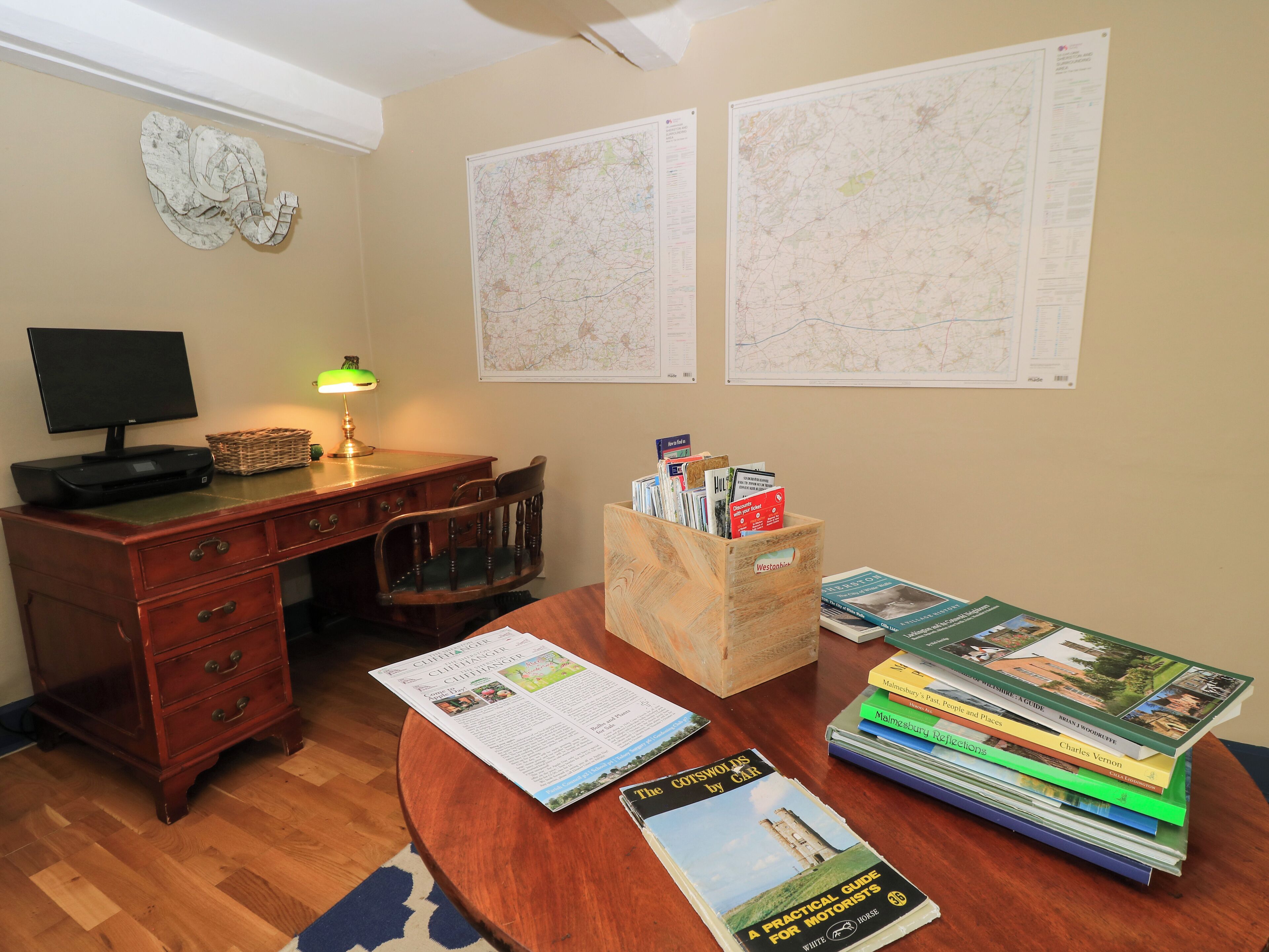 A room with a wooden desk, a computer, a lamp, a chair, a map, and a stack of books.