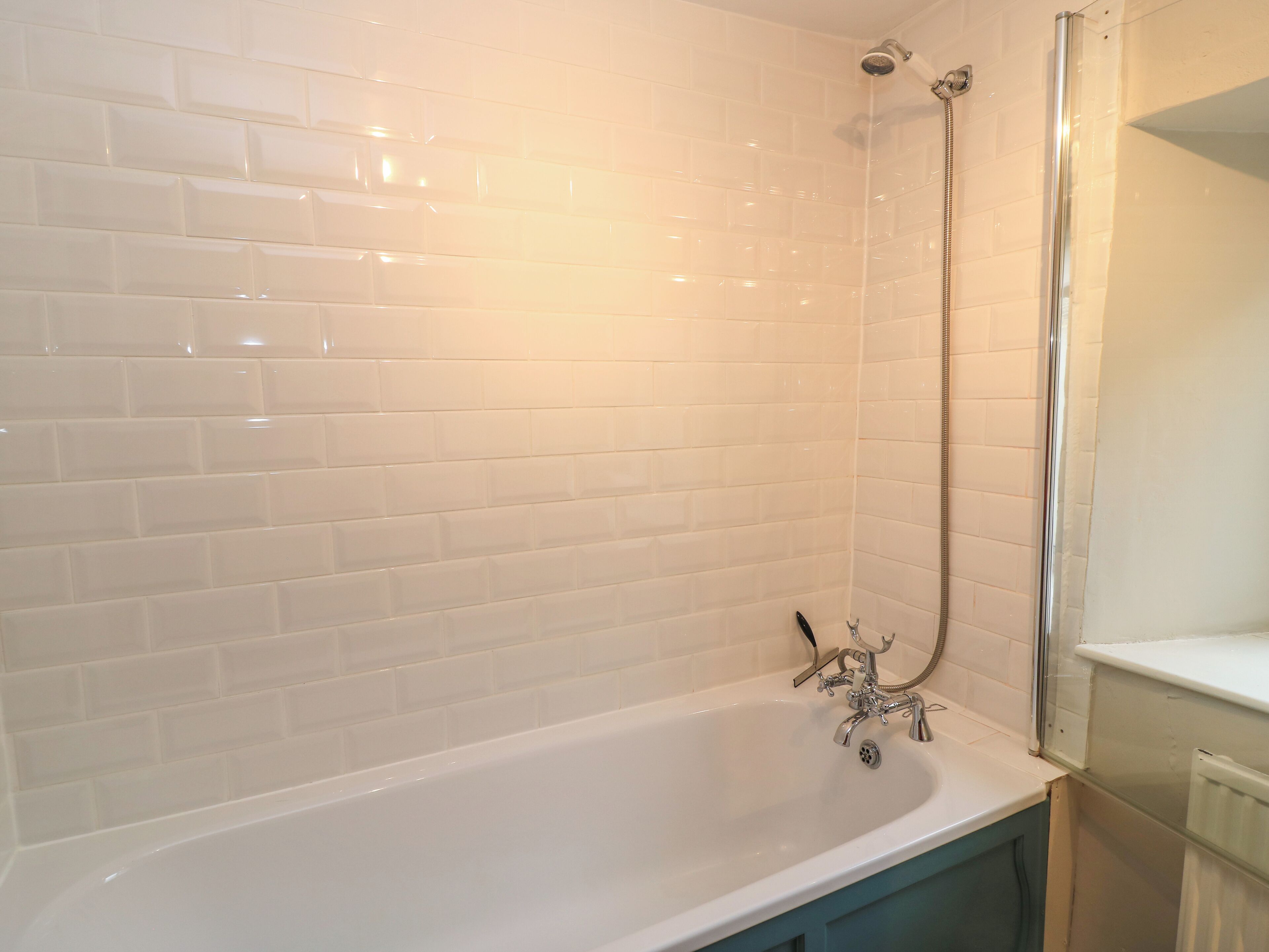 Une salle de bain avec des murs recouverts de carreaux blancs, une baignoire blanche et une douche dotée d’un robinet chromé.