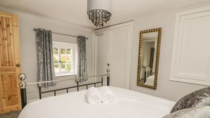 A bedroom with a bed, two towels, a chandelier, a window with curtains, a mirror, and a wooden door. - Ploughman's (Diss)