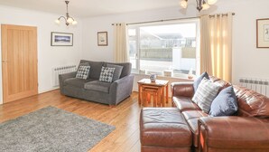 A living room with a grey sofa, a brown leather sofa, a wooden coffee table, and a grey rug. There are two windows with curtains, a wooden door, and a radiator. - Trem Y Garn (Pwllheli)