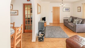 A living room with a wooden floor, a grey sofa, a brown leather ottoman, a grey rug, and a wooden door with glass panels. - Trem Y Garn (Pwllheli)