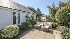 A patio with a sofa and a table, surrounded by greenery and a house with a white exterior. - Trem Y Garn (Pwllheli)