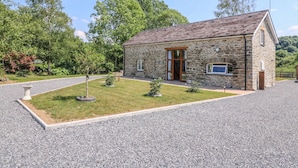 A stone building with a sloped roof, a front door, and a small garden area with a tree and a decorative planter.