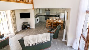 A spacious living area with a high ceiling, a dining table set for six, a kitchen with modern appliances, and a cozy seating area with a green sofa and a TV mounted on the wall.