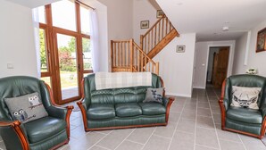 A spacious living room with a green leather sofa, two matching armchairs, a wooden staircase, and a large window offering a view of the outdoors.