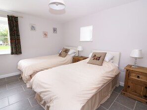 A bedroom with two beds, a window with curtains, a nightstand with a lamp, and a wooden cabinet. - Cilgwyn Bach (Newport)