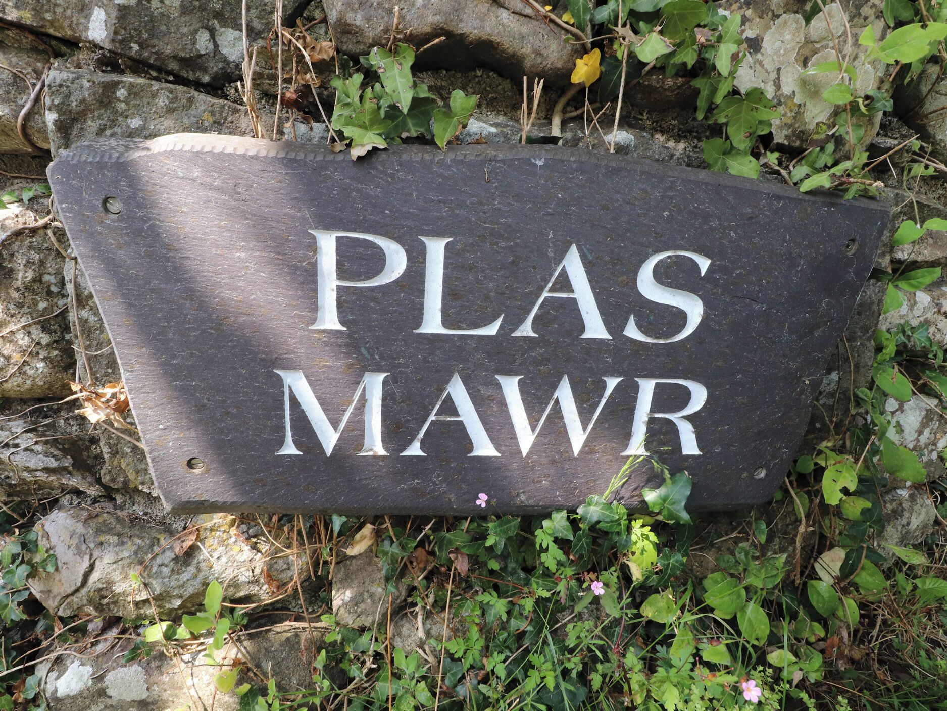A stone sign with the inscription 'PLAS MAWR' surrounded by greenery and a stone wall.