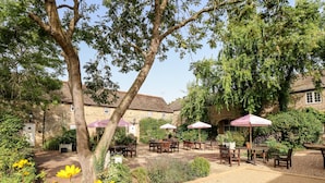 An outdoor patio area with tables and chairs, surrounded by greenery and a stone building. - Seaton (Oakham)