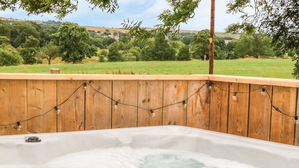 Un jacuzzi con terraza de madera y luces colgantes, rodeado de un paisaje verde exuberante.