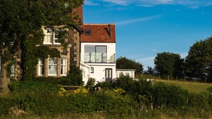 A two-story house with a large window and a balcony, surrounded by greenery and a field. - Emblestones (Alnwick)