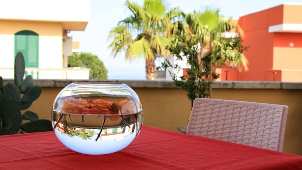 A fishbowl on a table with a red tablecloth, surrounded by a building with a green door and palm trees in the background. - B&B Barone (Castro)