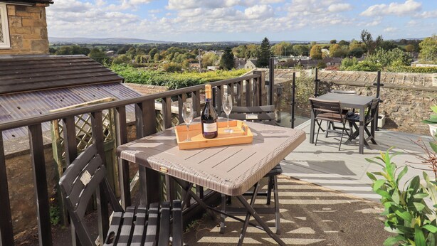 Eine Terrasse im Freien mit einem Holztisch und Stühlen, einer Weinflasche und einem Glas. Die Terrasse bietet einen Blick auf eine malerische Landschaft mit Bäumen und Gebäuden in der Ferne.