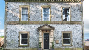 A two-story stone building with a central entrance, flanked by windows on each floor, and a small garden area with potted plants. - Magistrates Chambers (Carnforth)