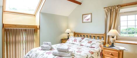 A bedroom with a bed, two bedside tables, a window with curtains, a wooden ceiling, and a skylight.