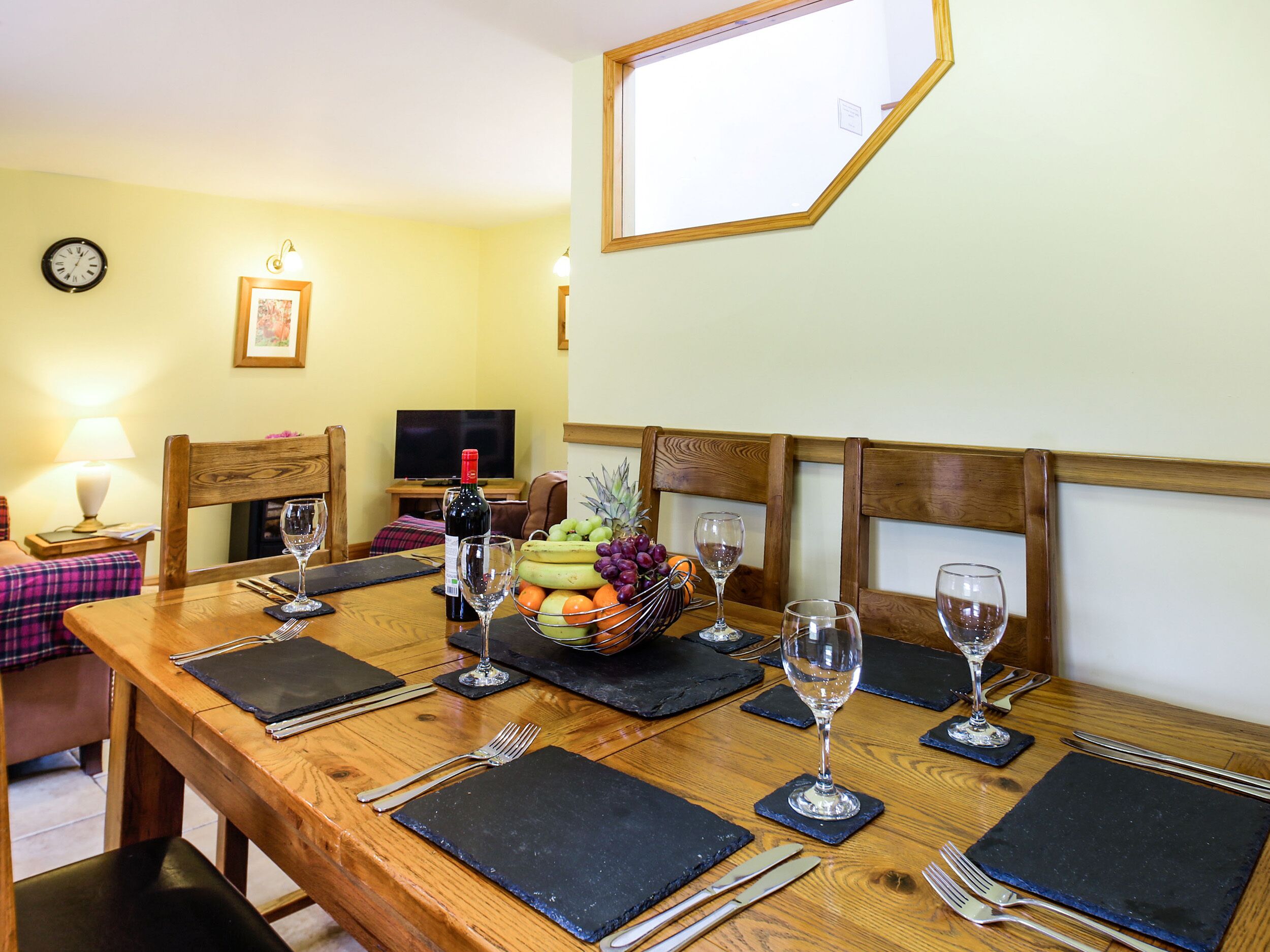 Una mesa de comedor lista con copas de vino, cubiertos y una cesta de fruta.
