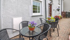 An outdoor seating area with round tables and wicker chairs, a white building with a red door, and flower planters. - Y Bwthyn (Tywyn)