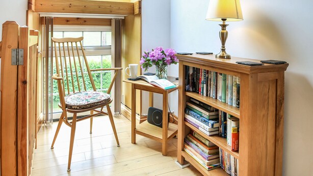A cozy reading nook with a wooden chair, a small table with a lamp, and a bookshelf filled with books. - Y Bwthyn (Tywyn)