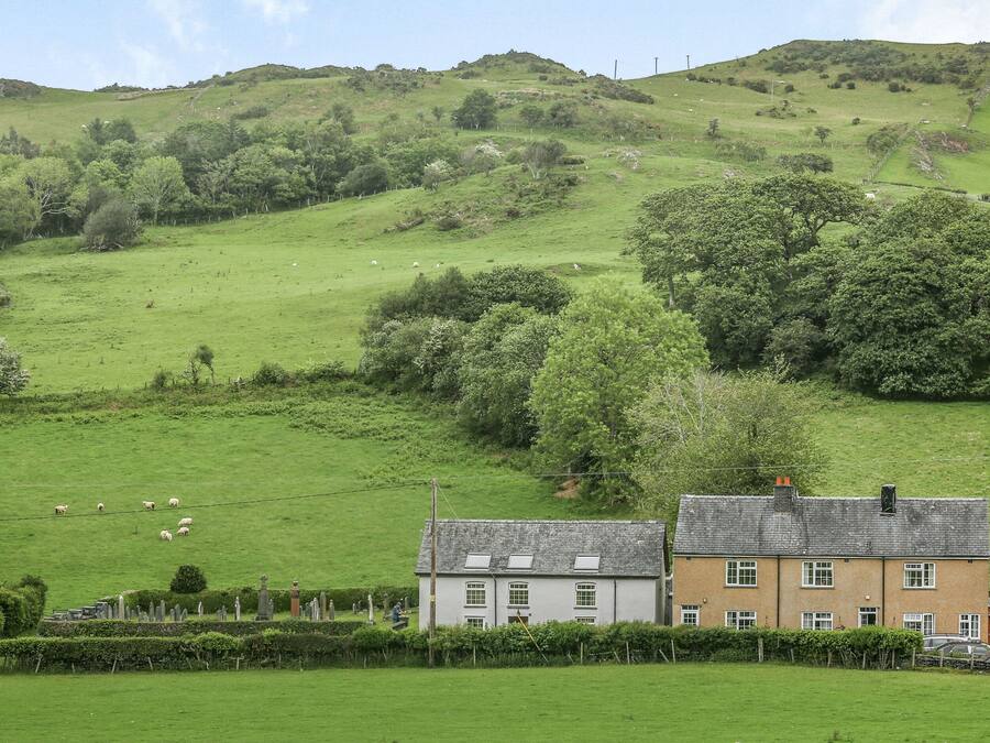 Un paysage rural avec une maison blanche et un bâtiment brun, entourés de champs verdoyants et d’une colline en arrière-plan.