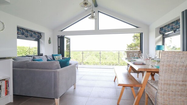 En rummelig opholdsstue med en grå sofa, et spisebord med stole og en balkon med udsigt over grønne områder.