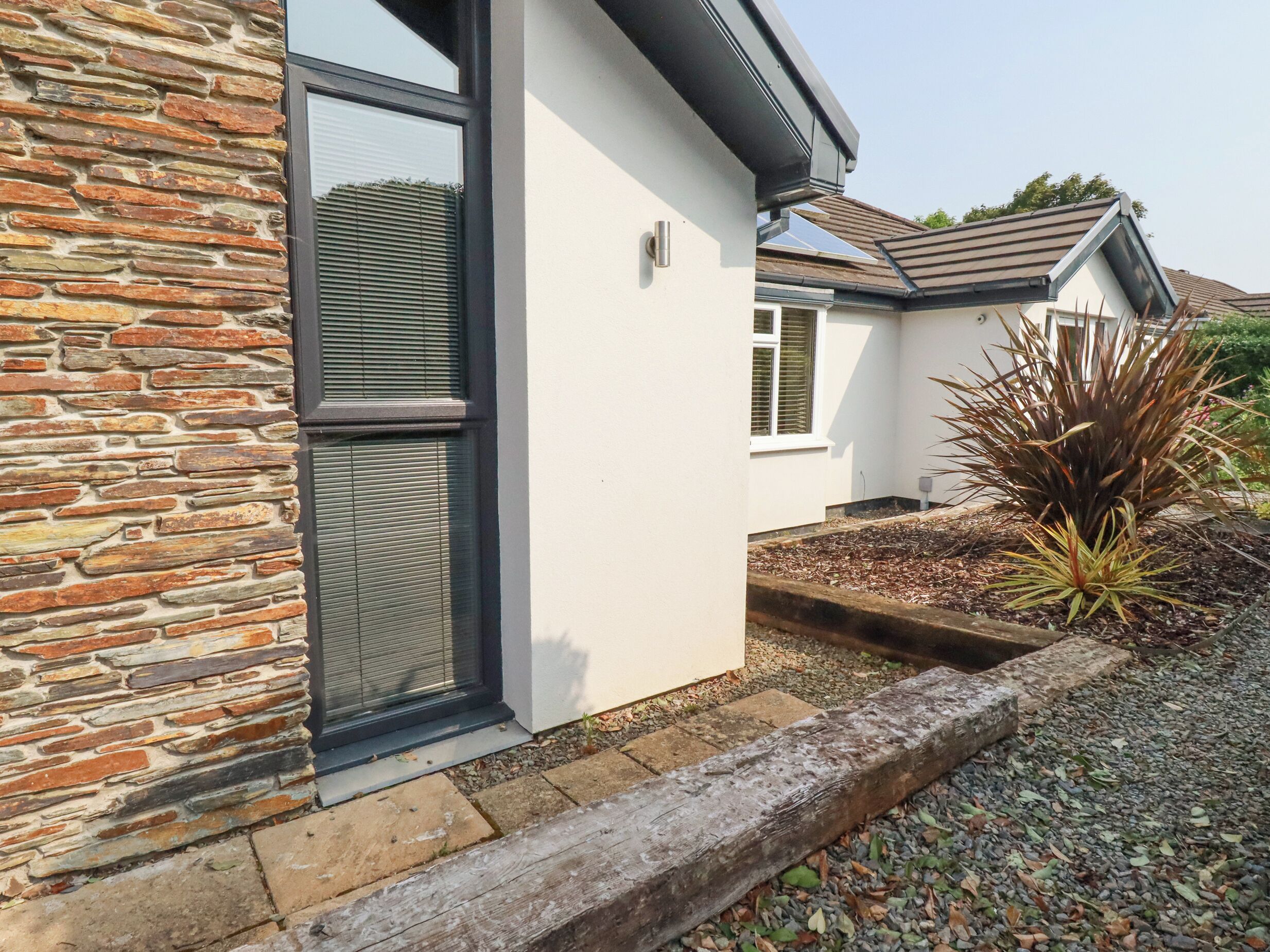 A modern house with stone and white exterior walls, a large window with blinds, and a small garden with a plant.