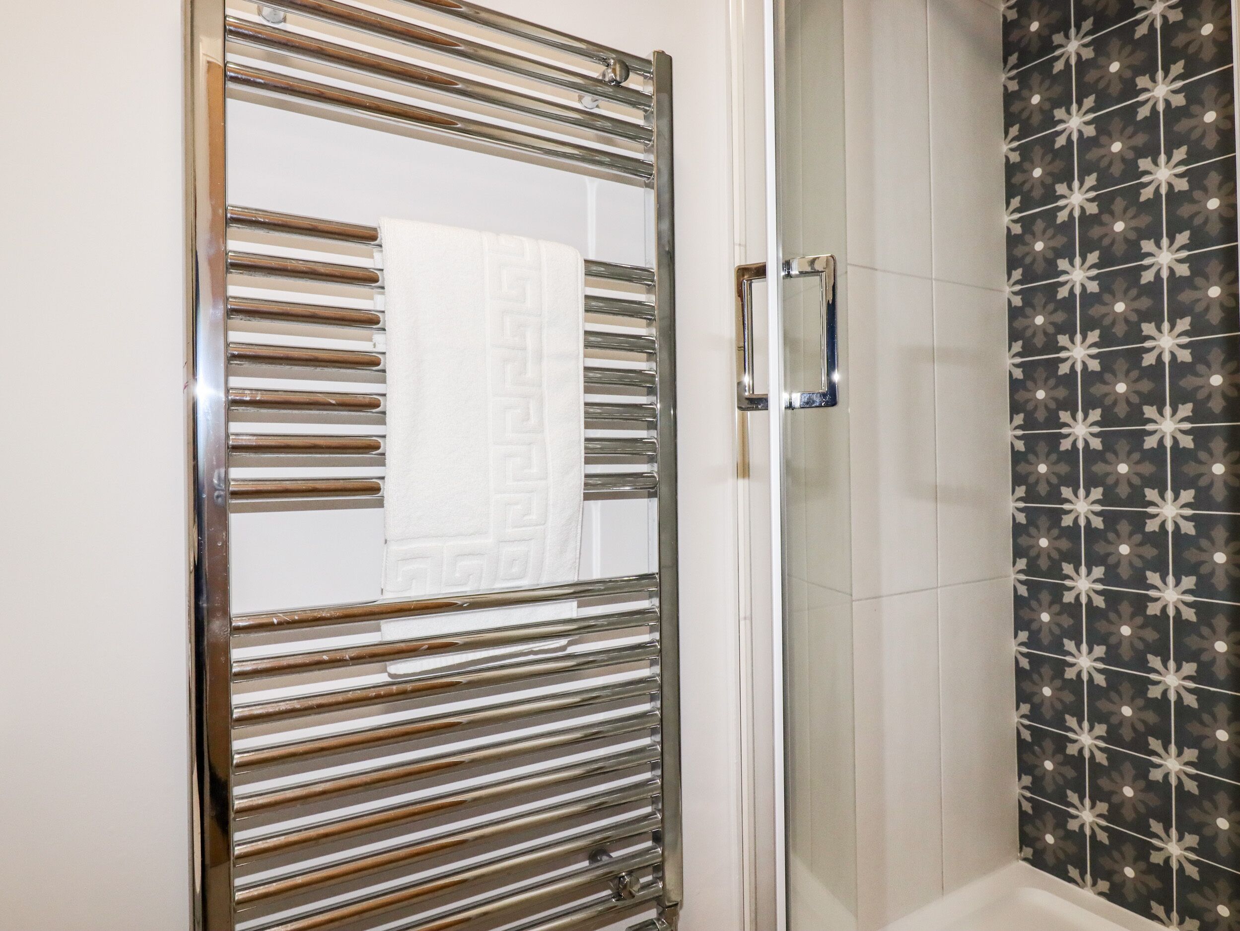 Een badkamer met een douchecabine van glas, een handdoek met een geometrisch patroon en een betegelde muur met een bloemenmotief.
