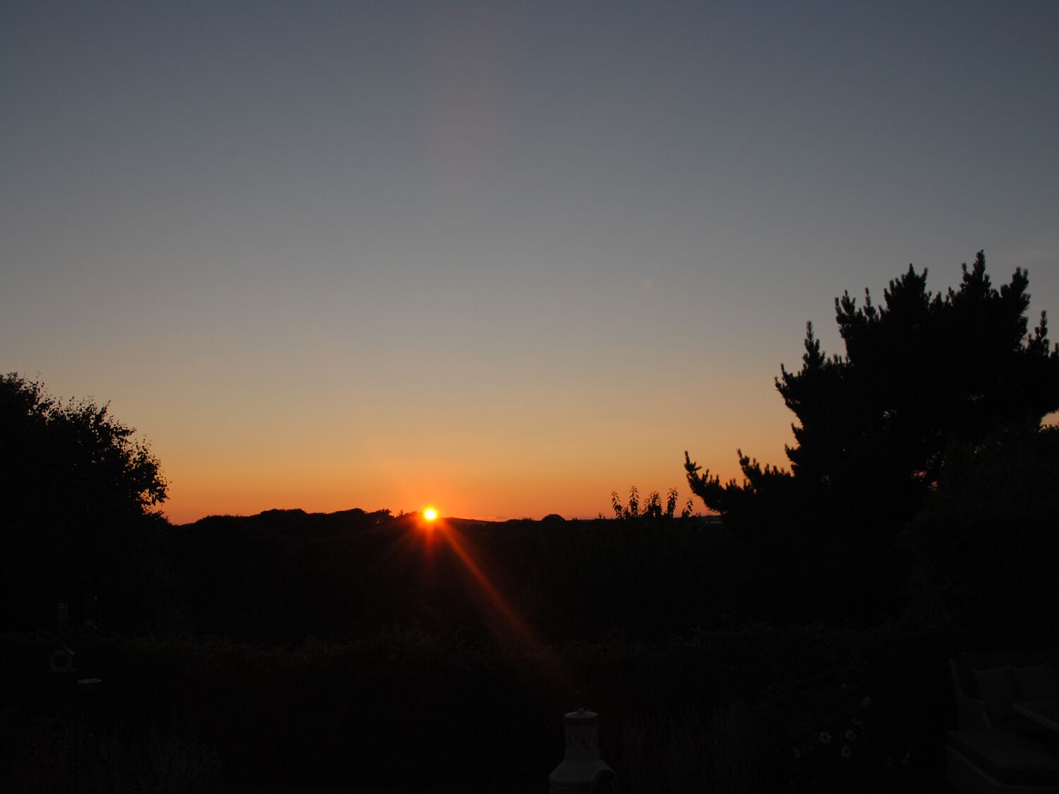 Um pôr do sol com o sol baixo no horizonte, árvores silhuetadas e céu claro.