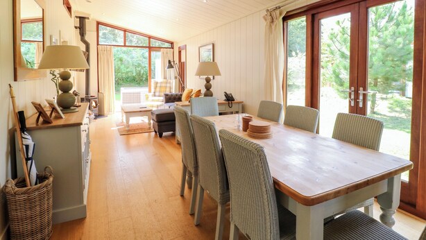 Un comedor amplio con una mesa y sillas de madera, ventanas grandes que ofrecen vistas a la vegetación y un rincón de descanso con sofá y sillones.