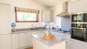 A modern kitchen with white cabinetry, a central island, a built-in oven, and a window with a patterned valance. - Brooke (Oakham)