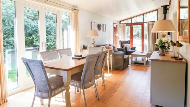 A spacious room with a wooden dining table, wicker chairs, a sofa, and a small table with a lamp. Large windows provide natural light and a view of greenery outside. - Brooke (Oakham)