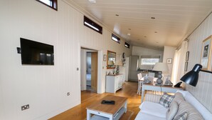Uma sala de estar espaçosa com sofá, mesa de centro e televisão na parede. Ao fundo, há uma área de cozinha com mesa e cadeiras.