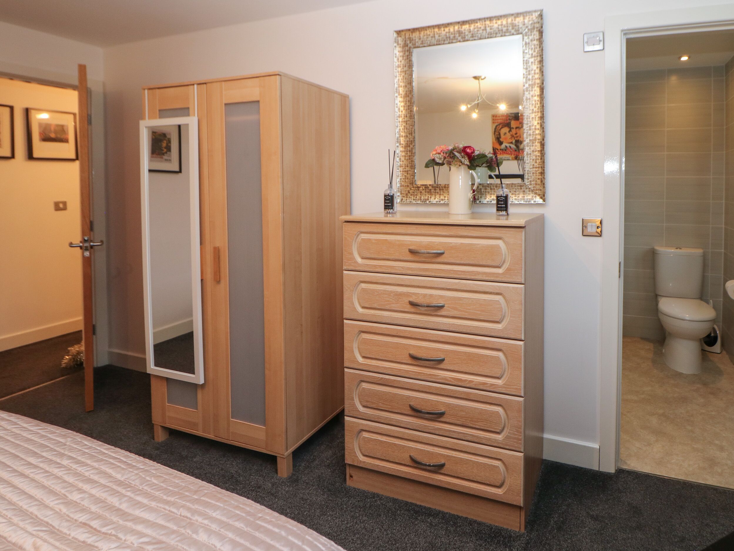 A bedroom with a bed, a wooden wardrobe, a dresser, a mirror, and a bathroom visible through an open door.