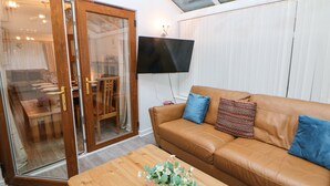 Un salon avec un canapé en cuir marron, une table basse en bois ornée de fleurs, un téléviseur fixé au mur et une porte-fenêtre coulissante donnant sur un coin repas.