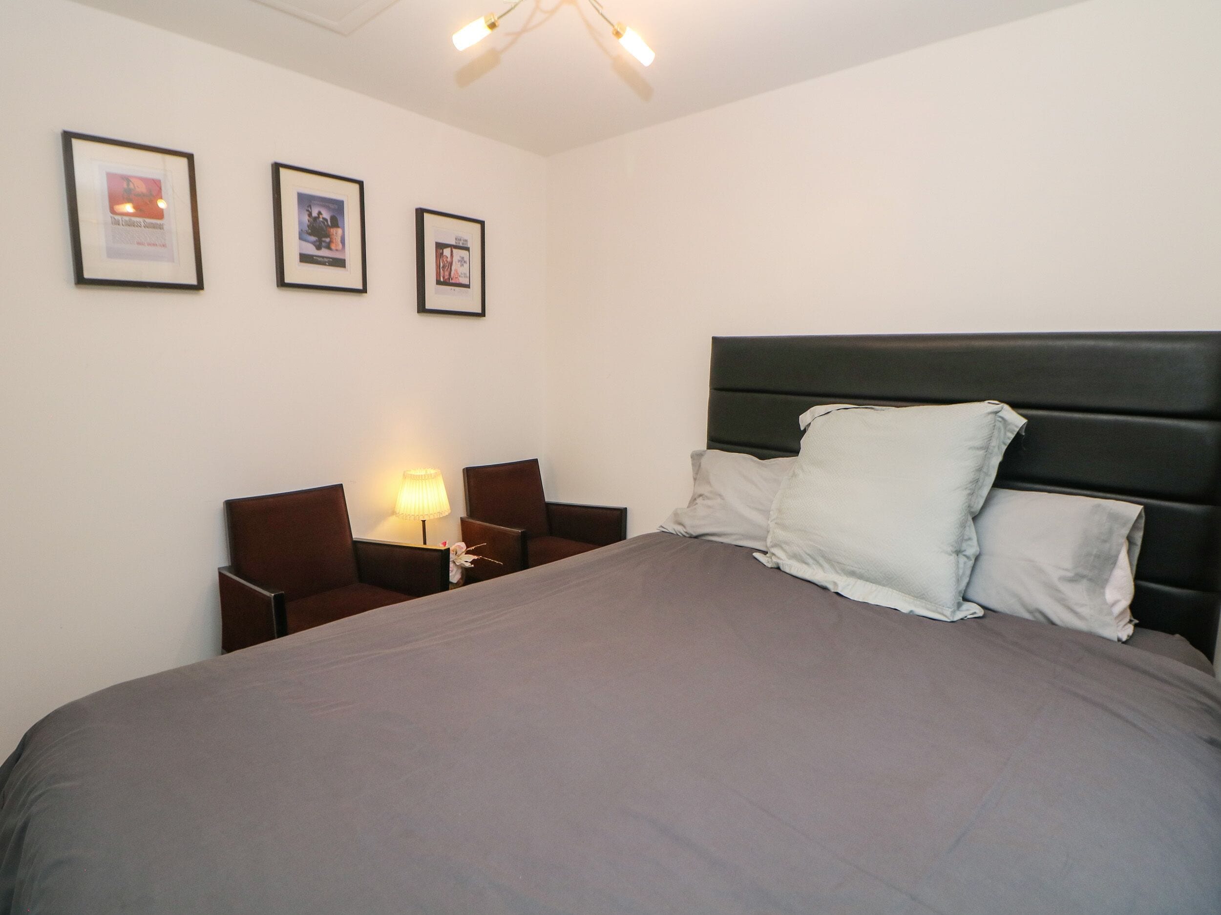 A neatly made bed with a grey bedspread and two pillows, flanked by two nightstands with lamps, in a room with white walls and framed pictures on the wall.