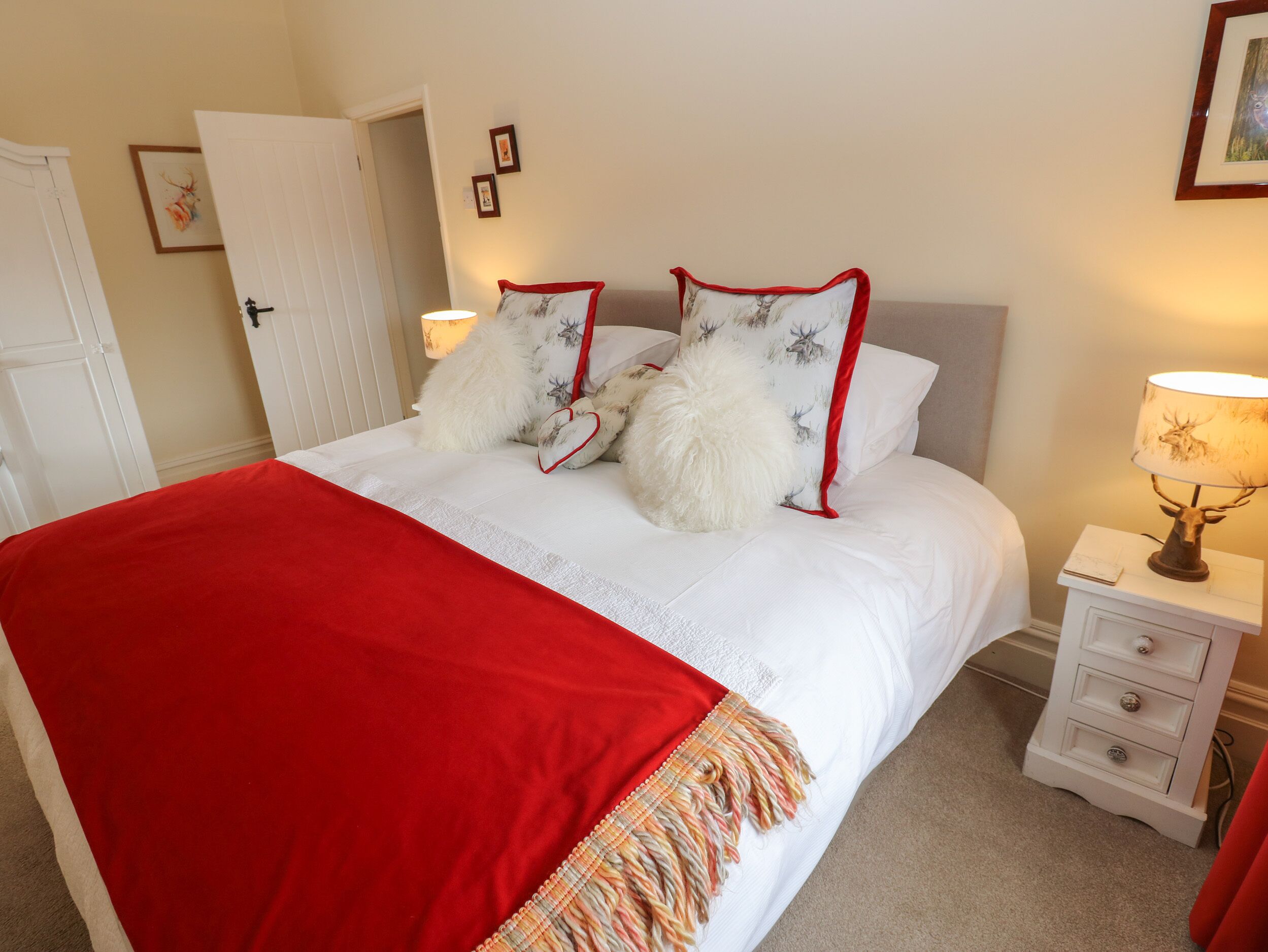 A neatly made bed with a red blanket, white pillows, and decorative pillows with reindeer prints. There is a bedside lamp and a small bedside table with drawers.