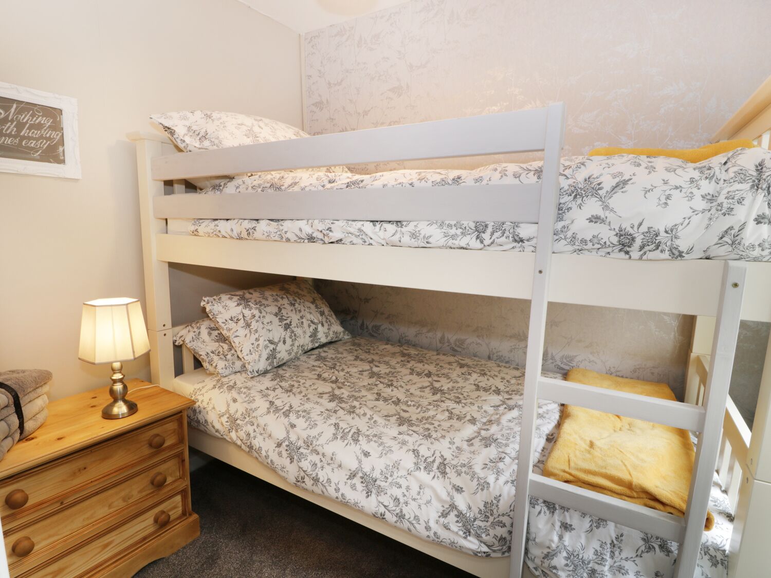 A bunk bed room with floral bedding, a bedside table with a lamp, and a small drawer unit.
