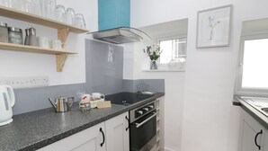 A modern kitchen with white cabinets, a black countertop, and a built-in oven. There are shelves with glassware and a window with a view of the outside. - Ty Coed (Caernarfon)