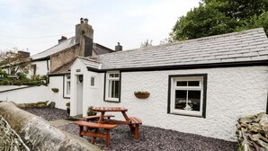 A white building with a stone wall, a wooden picnic table, and a window with a hanging plant. - Ty Coed (Caernarfon)