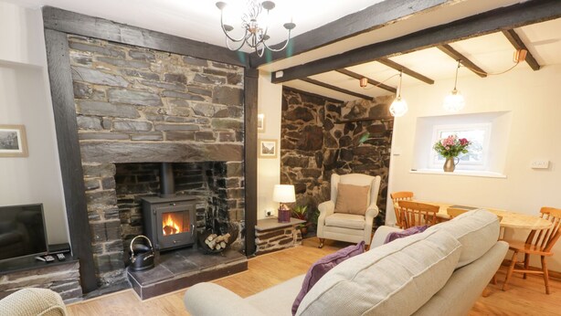 A cozy living room with a stone fireplace, a comfortable sofa, a dining area with wooden furniture, and a stone wall. - Ty Coed (Caernarfon)