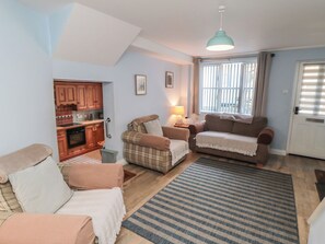 Una sala de estar acogedora con un sofá, un sillón, una mesita con una lámpara, y un área de cocina visible a través de una puerta abierta.