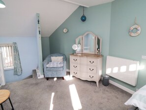 A bedroom with a sloped ceiling, a dresser with drawers, a chair, a radiator, and a window with curtains. - Bramla (Saltburn-by-the-Sea)