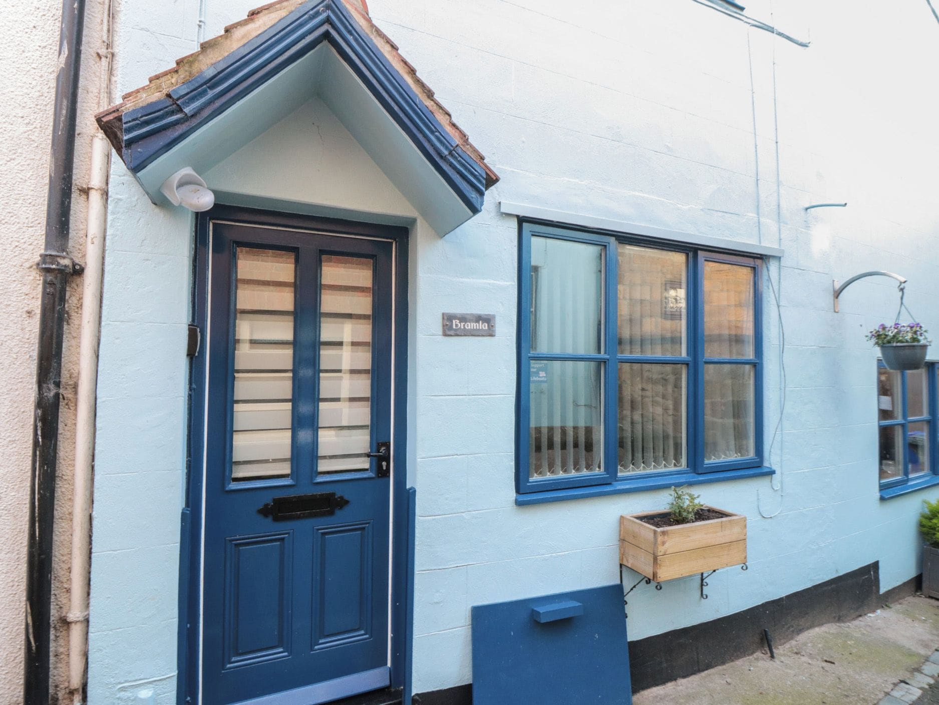 A blue-painted building with a door and windows, a nameplate reading 'Bramla', and a small wooden planter on the ground.