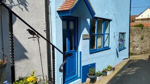 A blue cottage with a tiled roof, a black door, and multiple windows. There are potted plants and a metal railing visible. - Bramla (Saltburn-by-the-Sea)