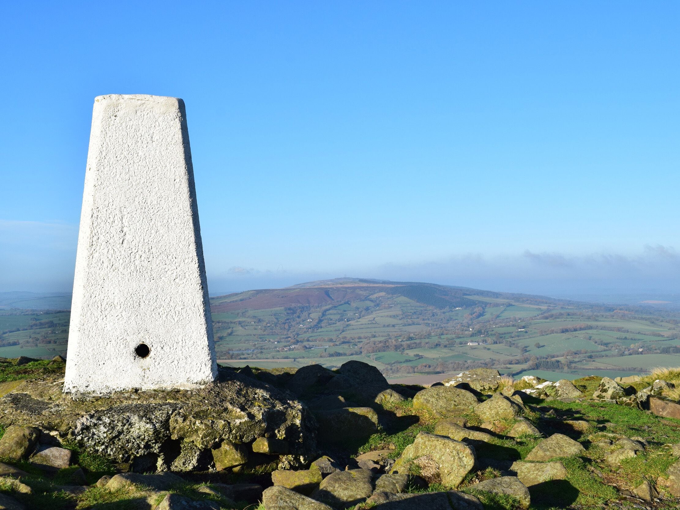 A stone cairn on a rocky outcrop with a panoramic view of rolling hills and distant fields.
