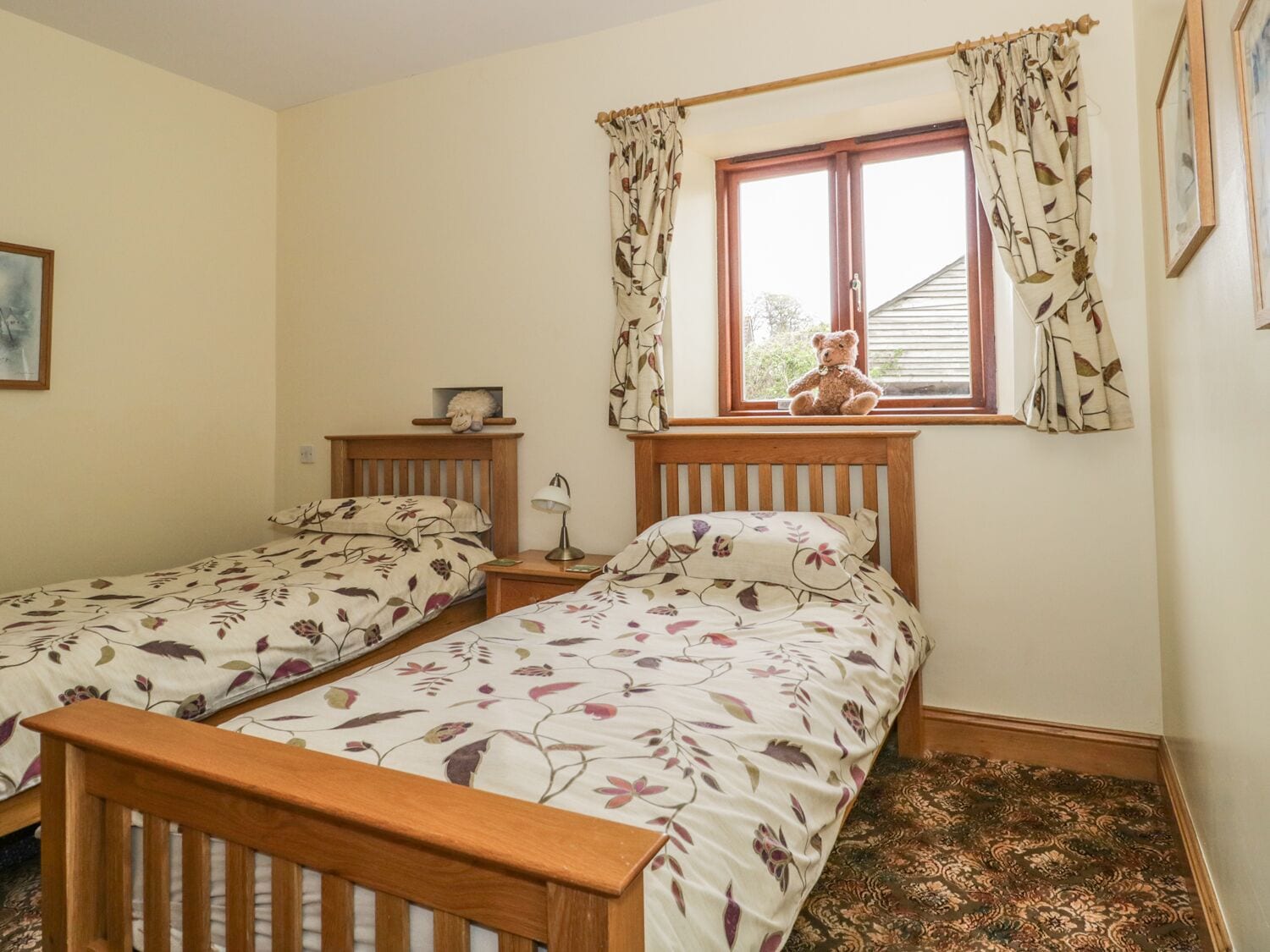A bedroom with two single beds, a window with curtains, a bedside lamp, and a teddy bear on one bed.